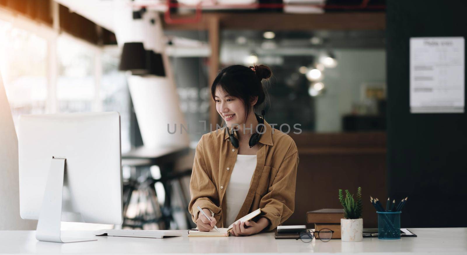 Asian woman wearing headphones using laptop and taking notes on homework that her teacher teaches at home