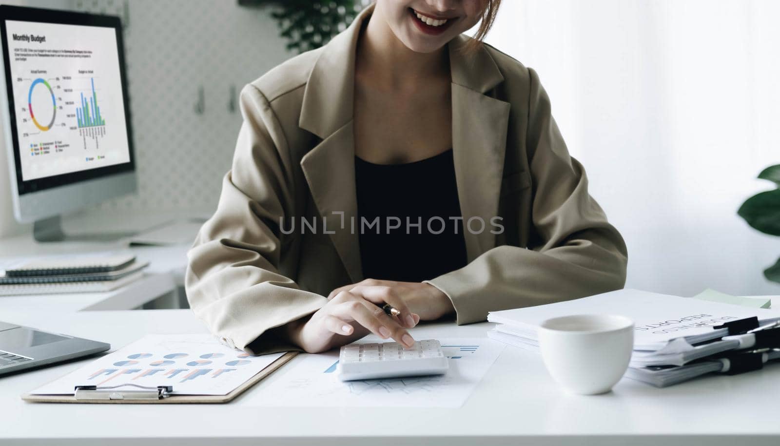 Smiling female accountant using calculator while sitting at office desk with digital tablet and financial documents..