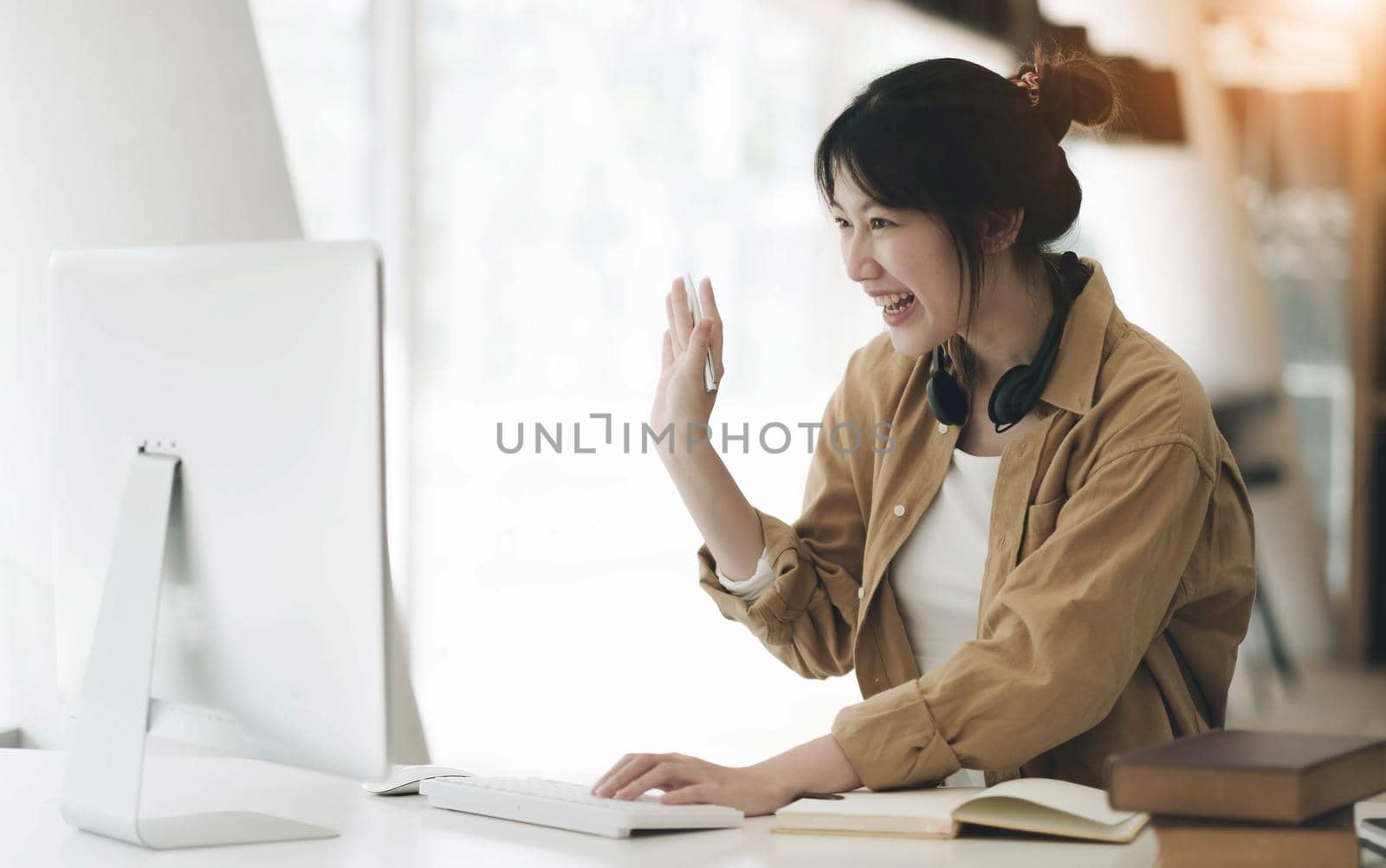 Head shot portrait smiling asian woman wearing headphones posing for photo at workplace, happy excited female wearing headset looking at laptop, sitting at desk with laptop, making video call by wichayada
