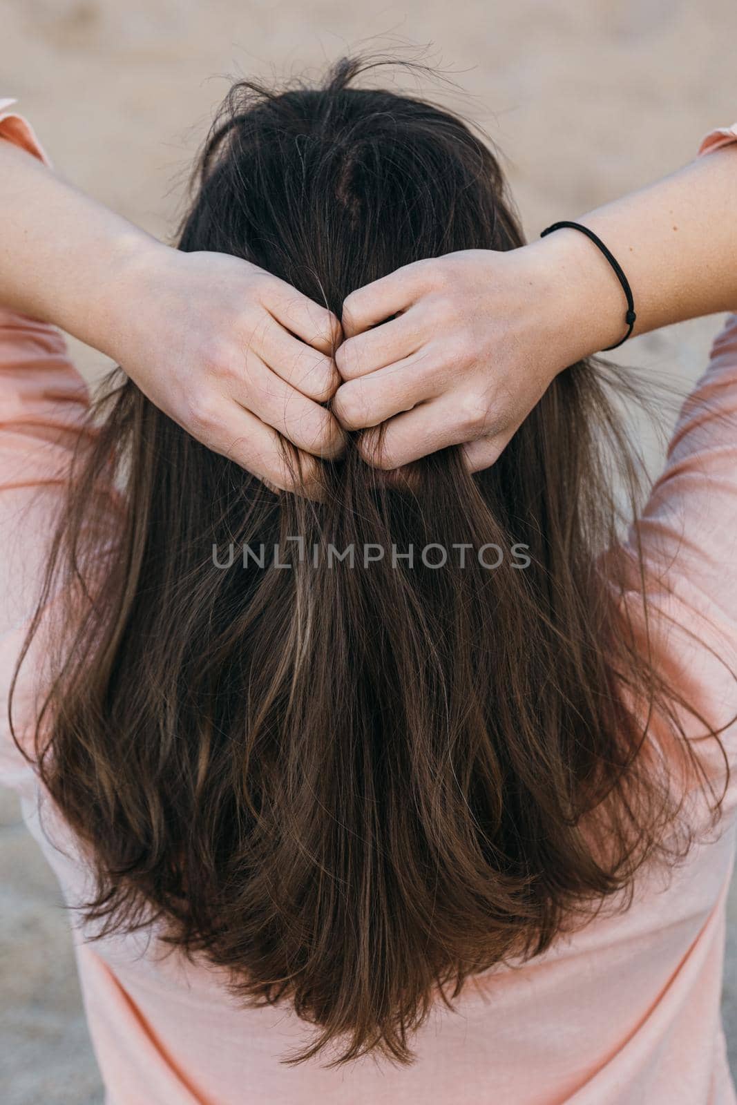A woman in pink shirt holding her natural hair with both hands. Brunette woman with long hair, view from the back