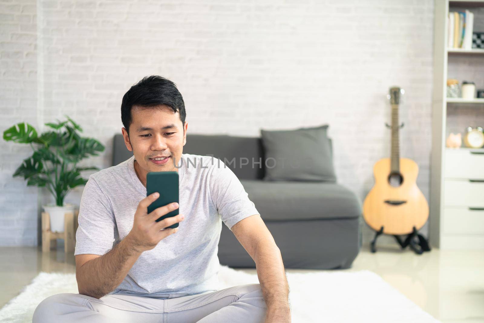 Smiling asian man wearing casual clothes sitting on a couch at the living room. Asian man using mobile phone chatting in living room. by Wmpix