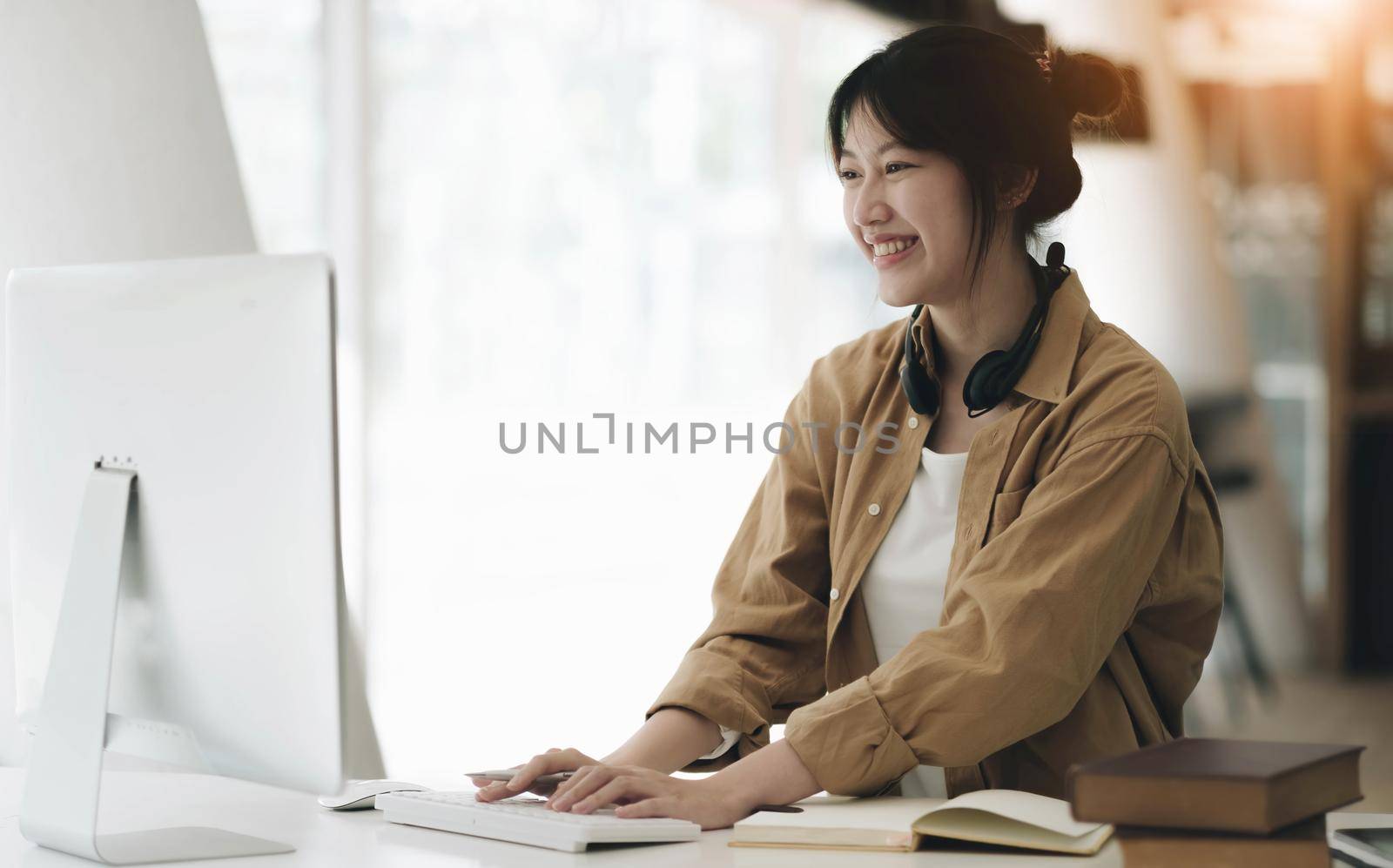 Head shot portrait smiling asian woman wearing headphones posing for photo at workplace, happy excited female wearing headset looking at laptop, sitting at desk with laptop, making video call.