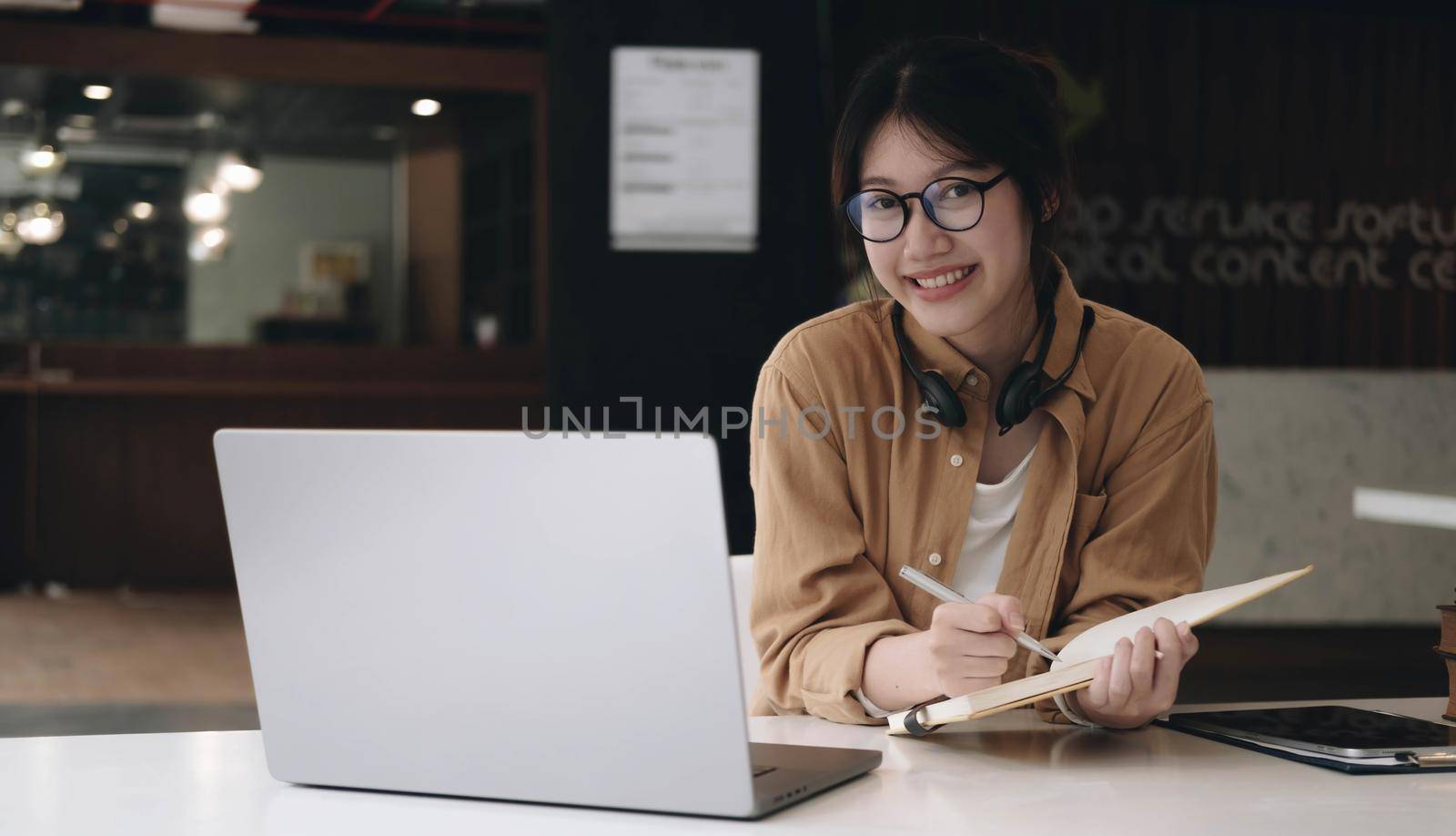 Happy asian woman wearing headset make notes talk by conference call doing video chat on laptop in office, smiling chinese support service agent write information consult customer by wichayada
