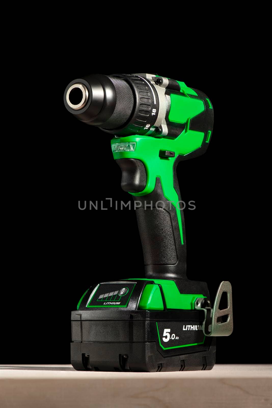 a cordless screwdriver stands on a wooden table on a black background. Cordless drill with lithium-ion battery in green. Professional tool for drilling holes and driving screws. by SERSOL