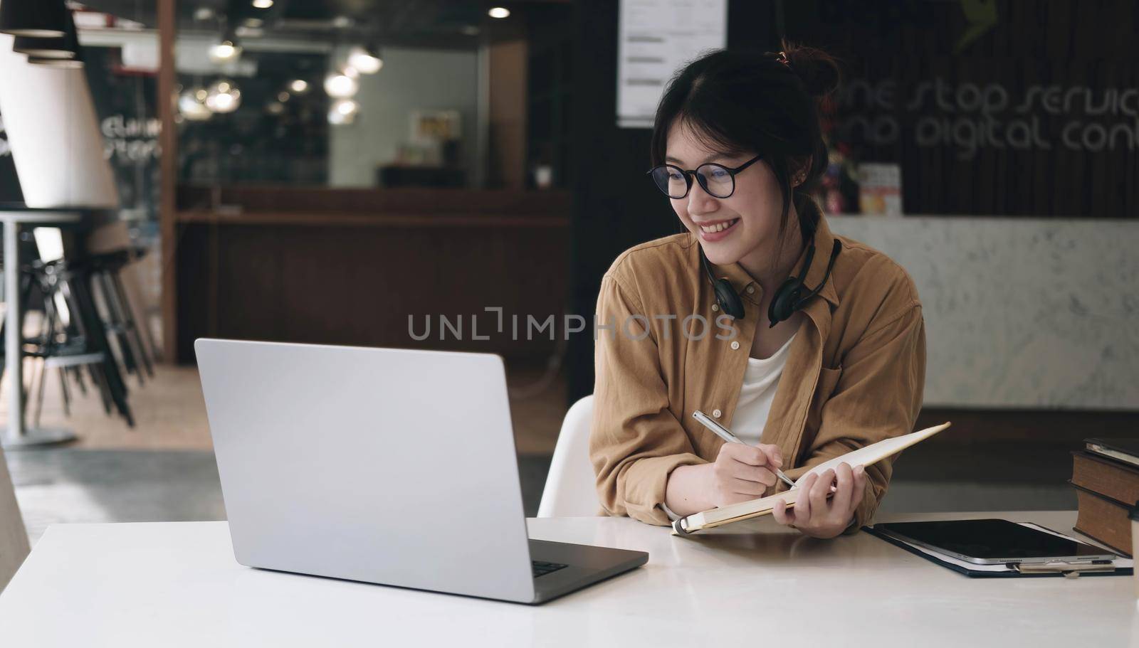 Happy asian businesswoman wearing headset make notes talk by conference call doing video chat on laptop in office, smiling chinese support service agent write information consult customer by wichayada