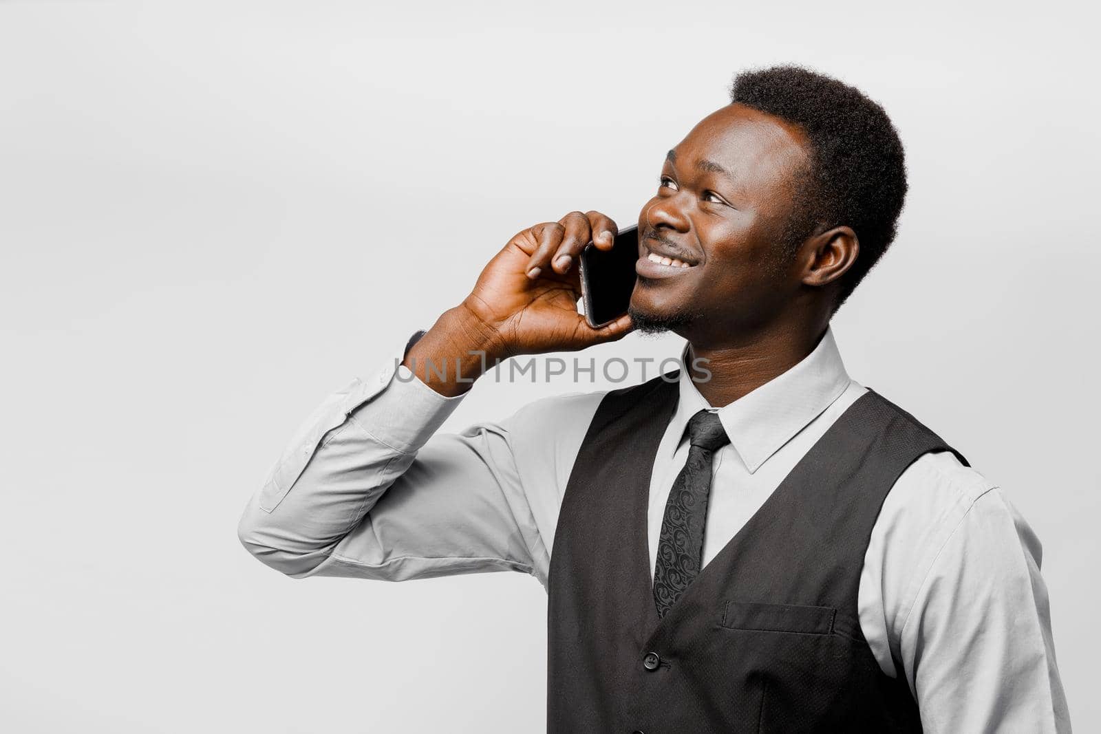 Happy black man speaks by phone and smiles. Smiling african man in dark vest, tie and white shirt.