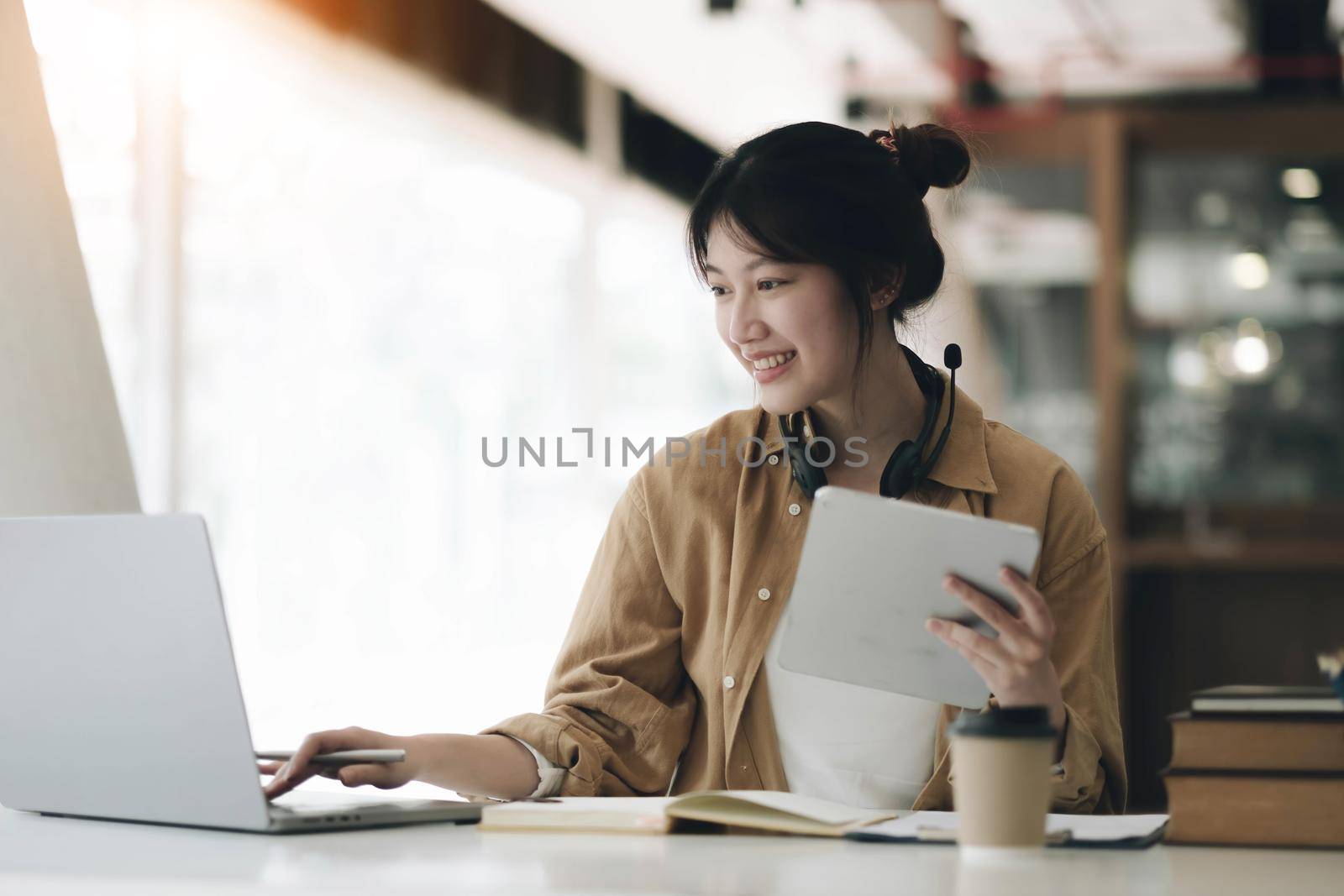 Asian woman wearing headphones around her neck using laptop and recording homework from teacher by wichayada