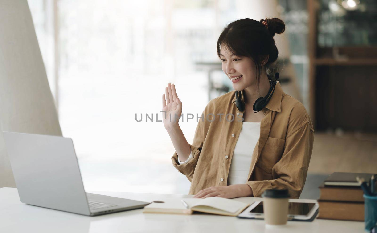 Asian woman wearing headphones on her neck using laptop and greeting friends on video call at home by wichayada