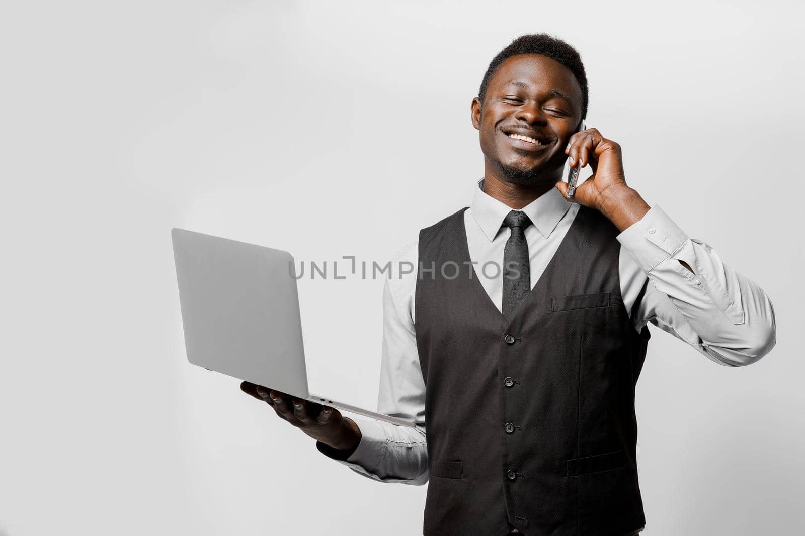Handsome black man speaks by phone and smiles. African with a laptop in hands. Speaking by phone in home at quarantine period coronavirus. by Rabizo