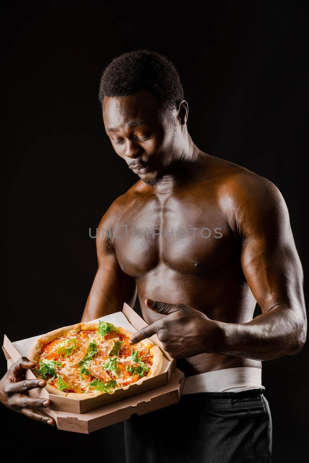 Surprised naked black man points pizza in cardboard box. Advertise for pizzeria. Isolated black backround. Discount for pizza.