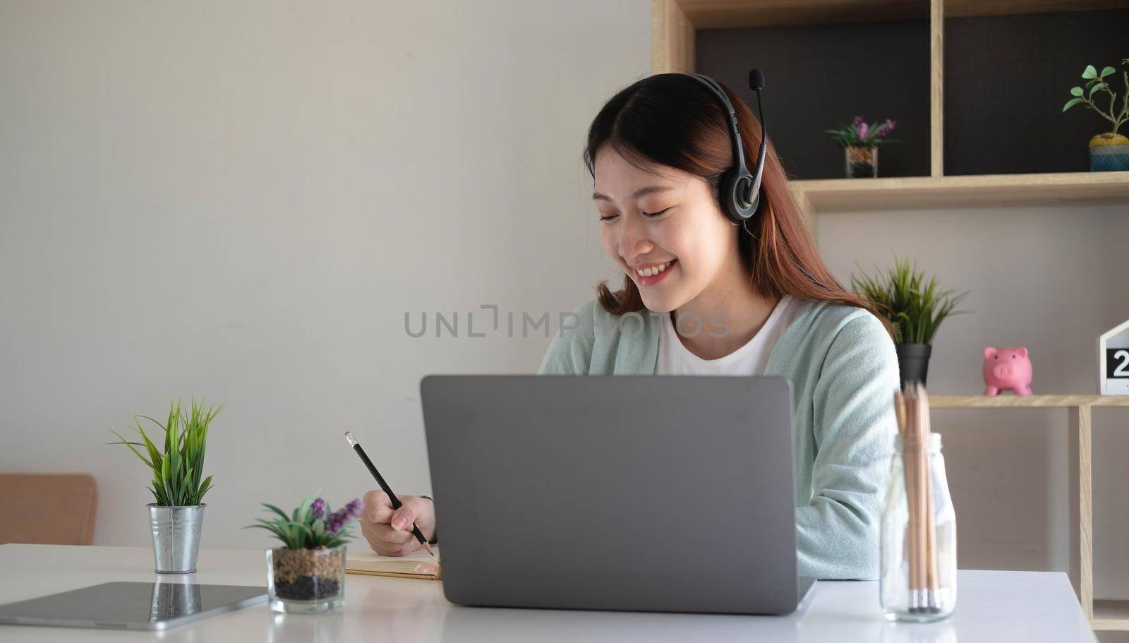 Asian schoolgirl in casual clothes is reading and doing homework video conference e-learning with teacher on laptop computer at home by wichayada