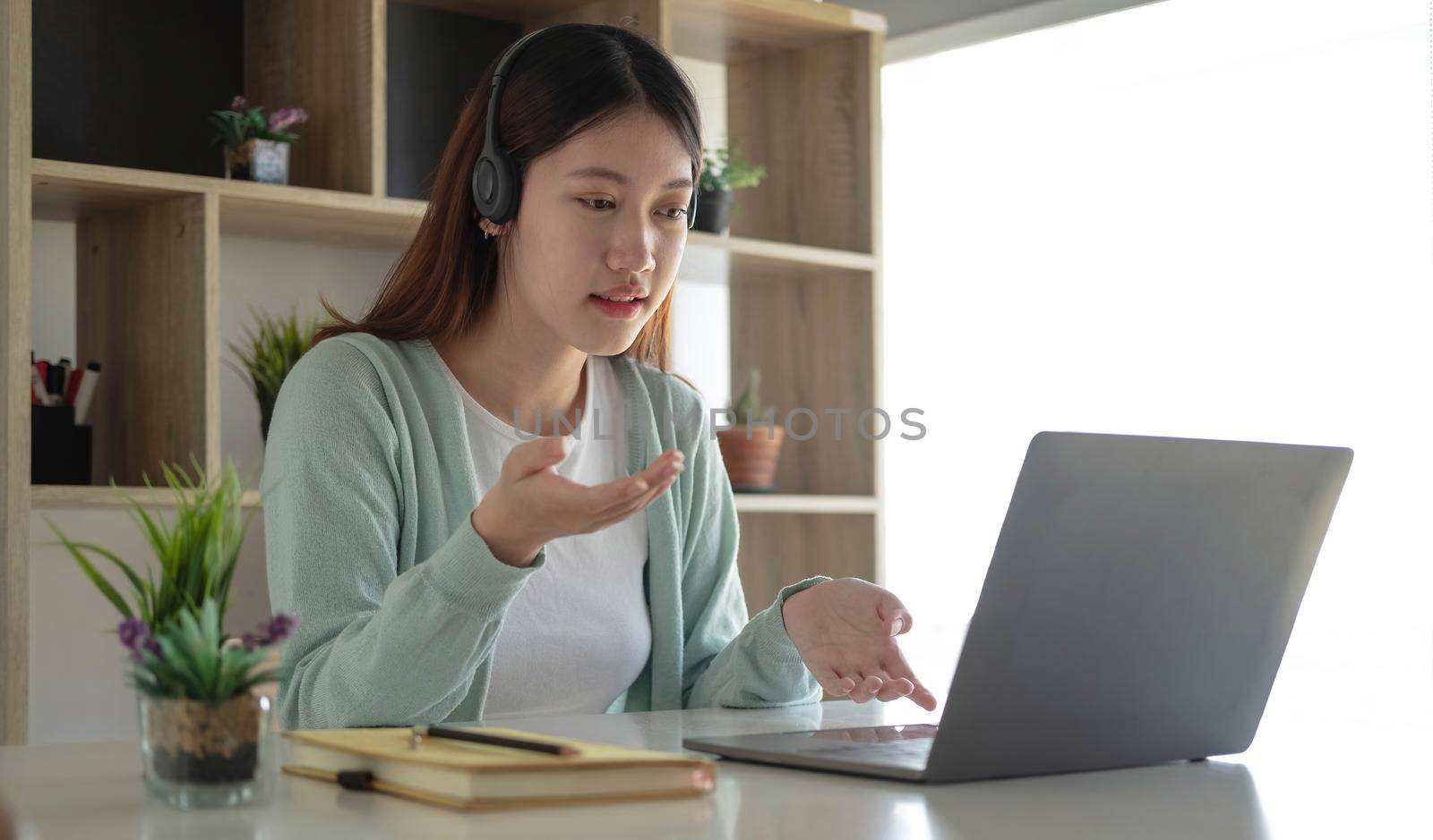 Happy young woman in headphones speaking looking at laptop making notes, girl student talking by video conference call, female teacher trainer tutoring by webcam, online training, e-coaching concept by wichayada