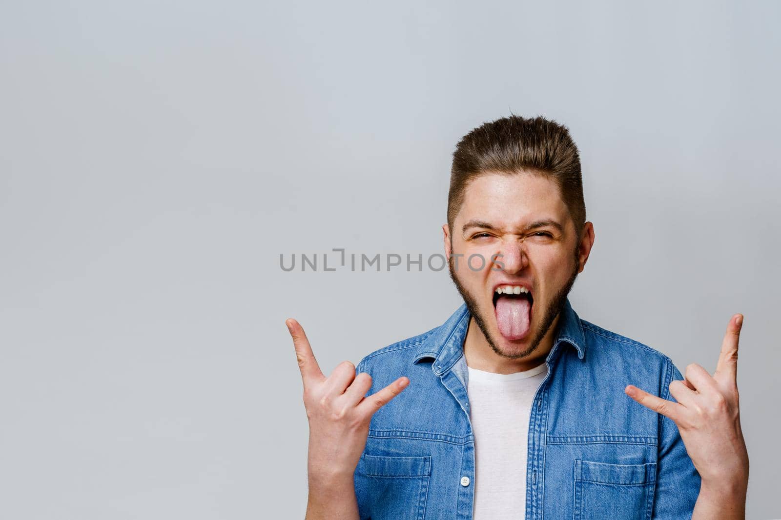 Confident bearded man shows tongue and makes rock n roll. Advertise for party. Young handsome man stants over white background and shows cool by Rabizo