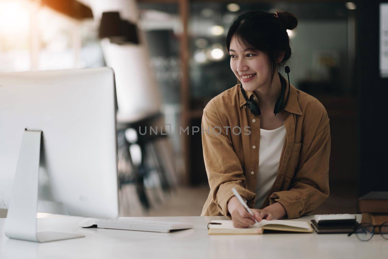 Asian woman wearing headphones using laptop and taking notes on homework that her teacher teaches at home