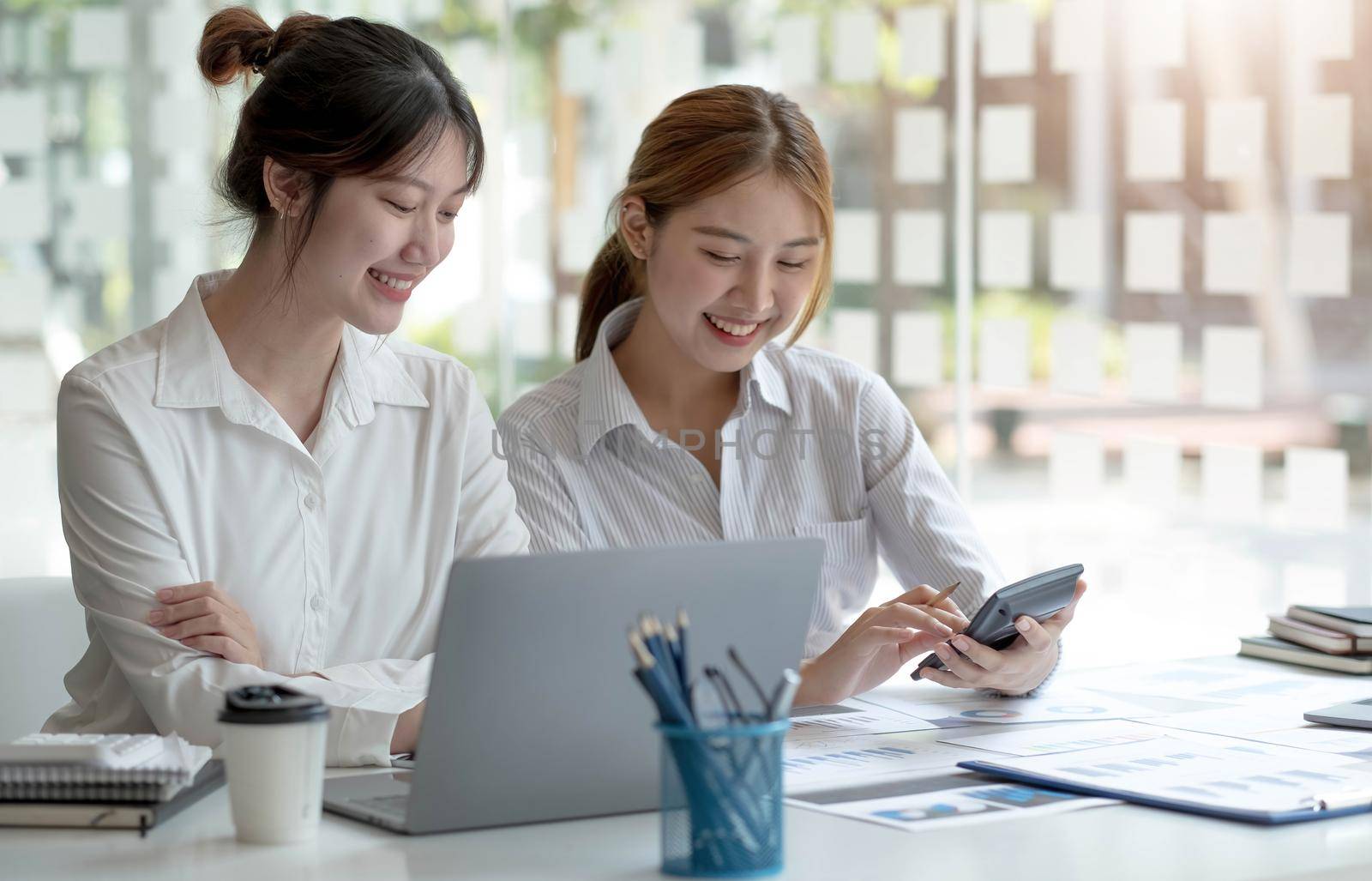 Two hardworking young women entrepreneurs working together on their laptop computers read screens with smiling faces in high angles..