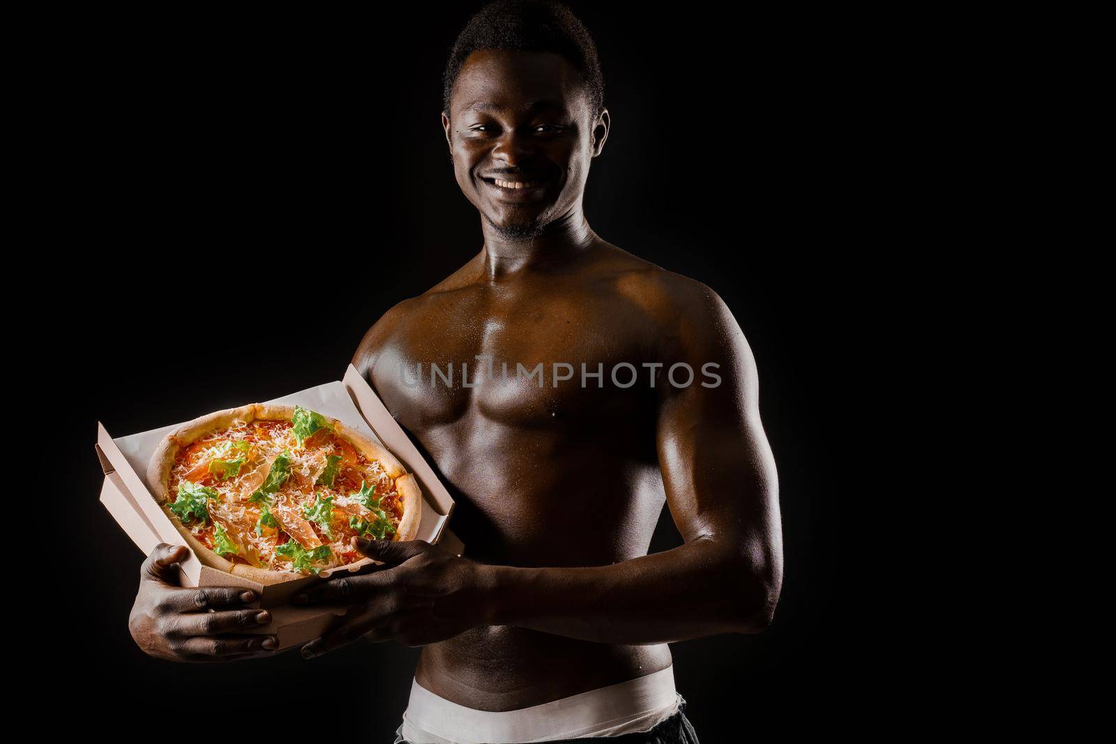 Funny black man with pizza smiles. Undressed african works in safety food delivery. Fit man with food from restaurand. Black undressed man with pizza. by Rabizo