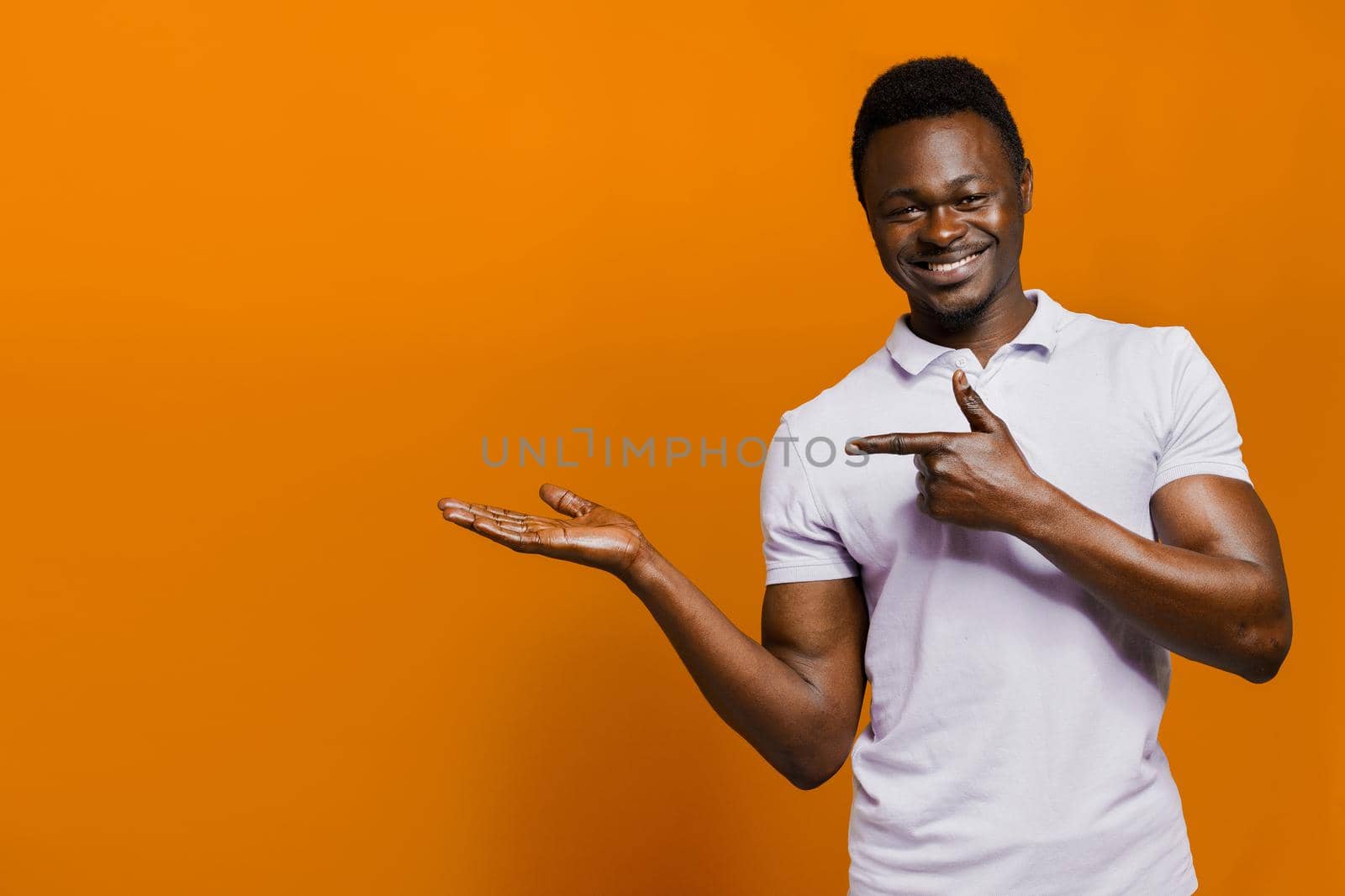 Happy black man smiles with white teeth and points to left side with empty space for advertise. African isolated orange background.