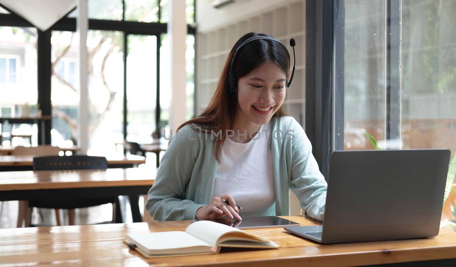 Happy young woman in headphones speaking looking at laptop making notes, girl student talking by video conference call, female teacher trainer tutoring by webcam, online training, e-coaching concept by wichayada