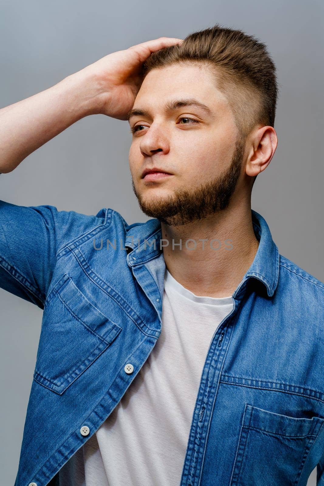 Young handsome man stands over white background and touches his hair after visiting barbershop. Confident man wears casual jeans shirt