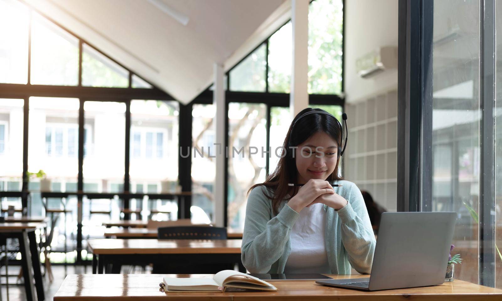 Smiling girl student wear wireless headphone study online with skype teacher, happy young woman learn language listen lecture watch webinar write notes look at laptop sit in cafe, distant education by wichayada