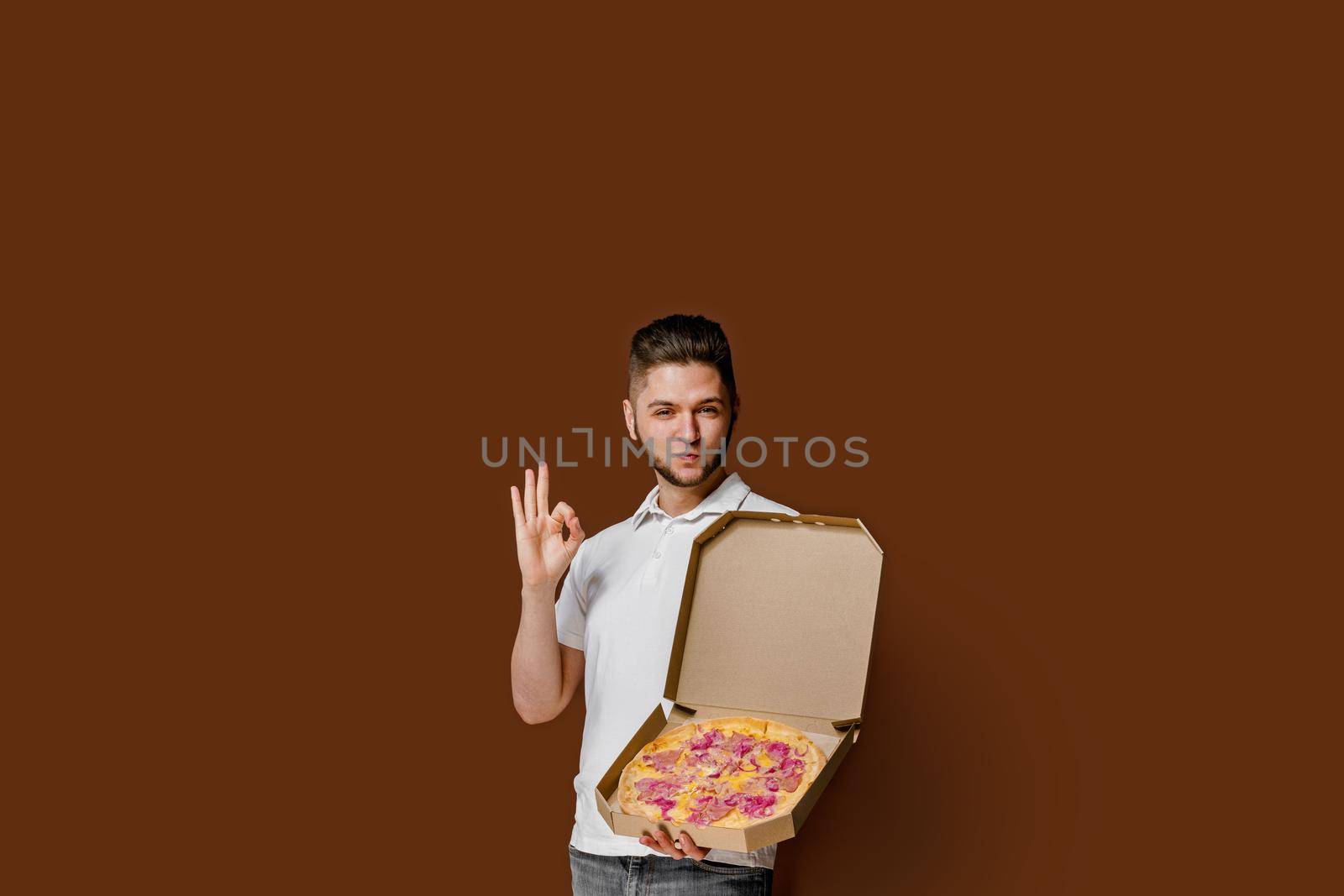 Online pizza safety delivery servise from restaurant. Young caucasian courier man shows ok and smile. Pizza with cheese boards advertise. by Rabizo