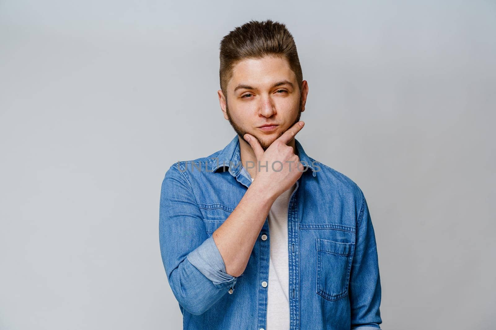 Young handsome man stands over white background and touches his beard after visiting barbershop. Confident man wears casual jeans shirt. by Rabizo