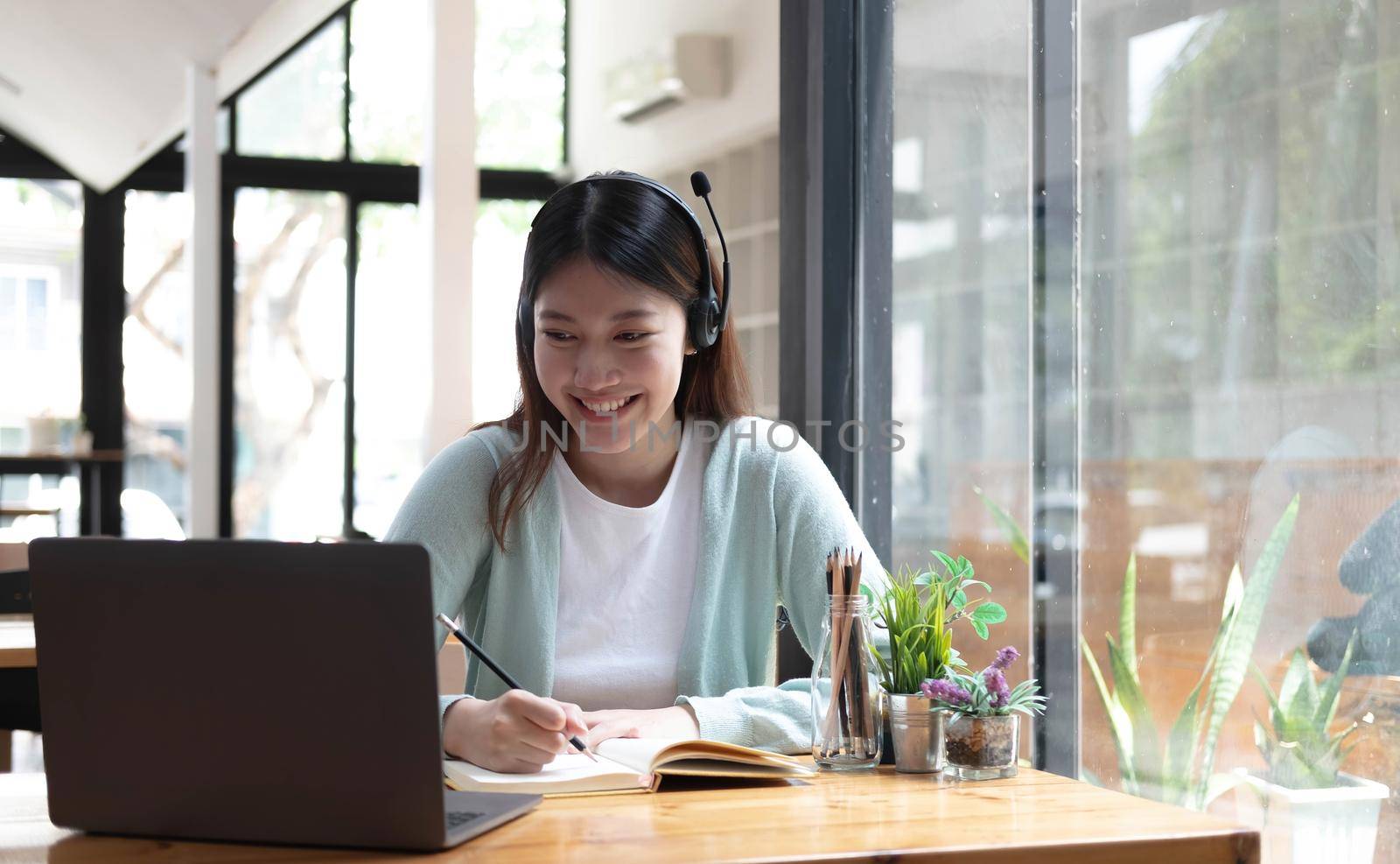 Smiling girl student wear wireless headphone study online with skype teacher, happy young woman learn language listen lecture watch webinar write notes look at laptop sit in cafe, distant education.