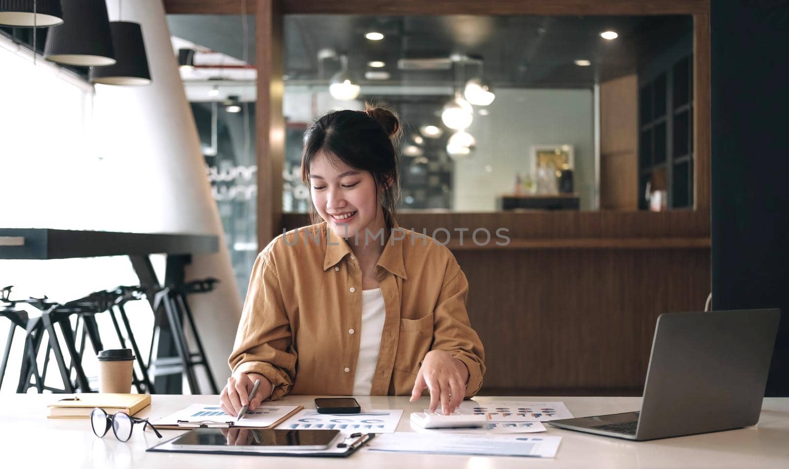 Happy young asian businesswoman sitting on her workplace and using calculator in the office. Young woman working at laptop in the office. by wichayada