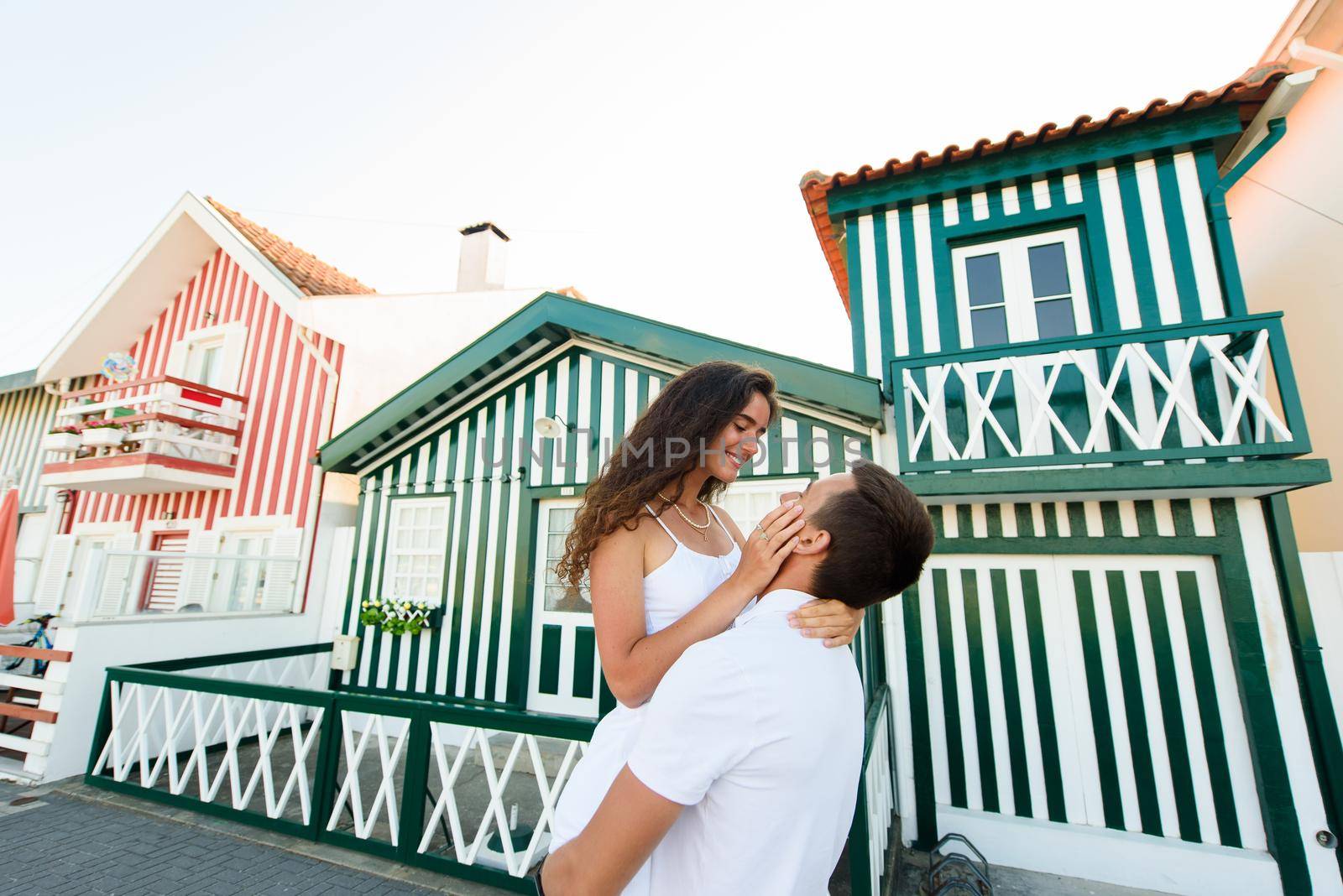 Handsome man puts up his girlfriend and kisses in Aveiro, Portugal near colourful and peaceful houses. Lifestyle of couple in love.
