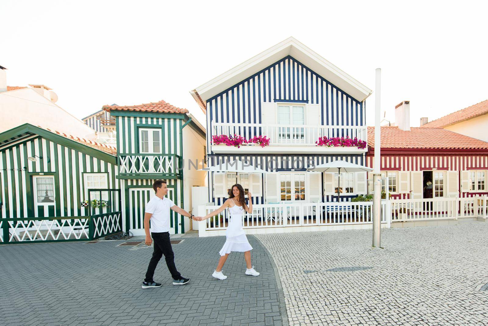 Young couple in white clothes walks around street in Aveiro, Portugal near colourful and peaceful houses. Lifestyle. Having fun, laughs, smiles. by Rabizo