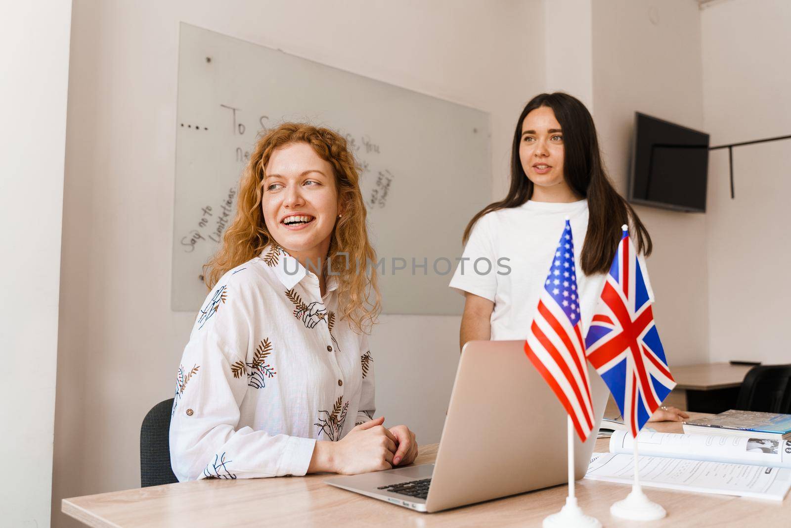 2 girls with laptop look at window in white class. Teacher of english asks student. Student answers to teacher. Working in group.