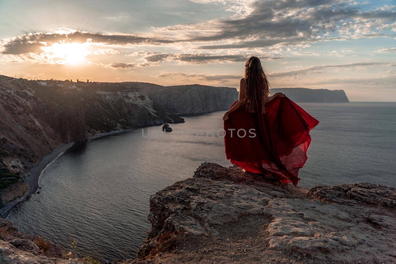 A girl with loose hair in a red dress waved her skirt on the yellow rocks overlooking the sea. In the background, the sun rises from behind the mountains