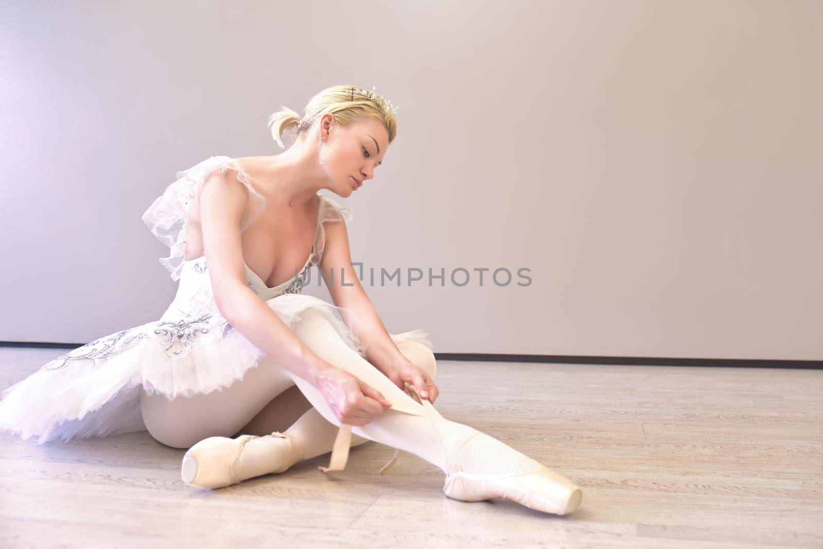 Photo of a tired ballerina sitting in a dance studio and puts on pointe shoes over beige wall background isolated