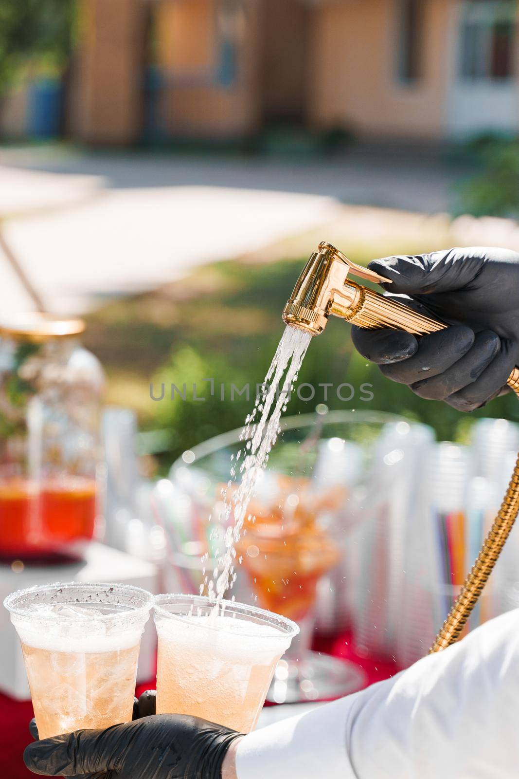 Waiter pours cider into plastic cups from bathroom faucet. Creative welcome drink zone. Bath with ice cubes, fruits, lemonades at hot summer day. by Rabizo
