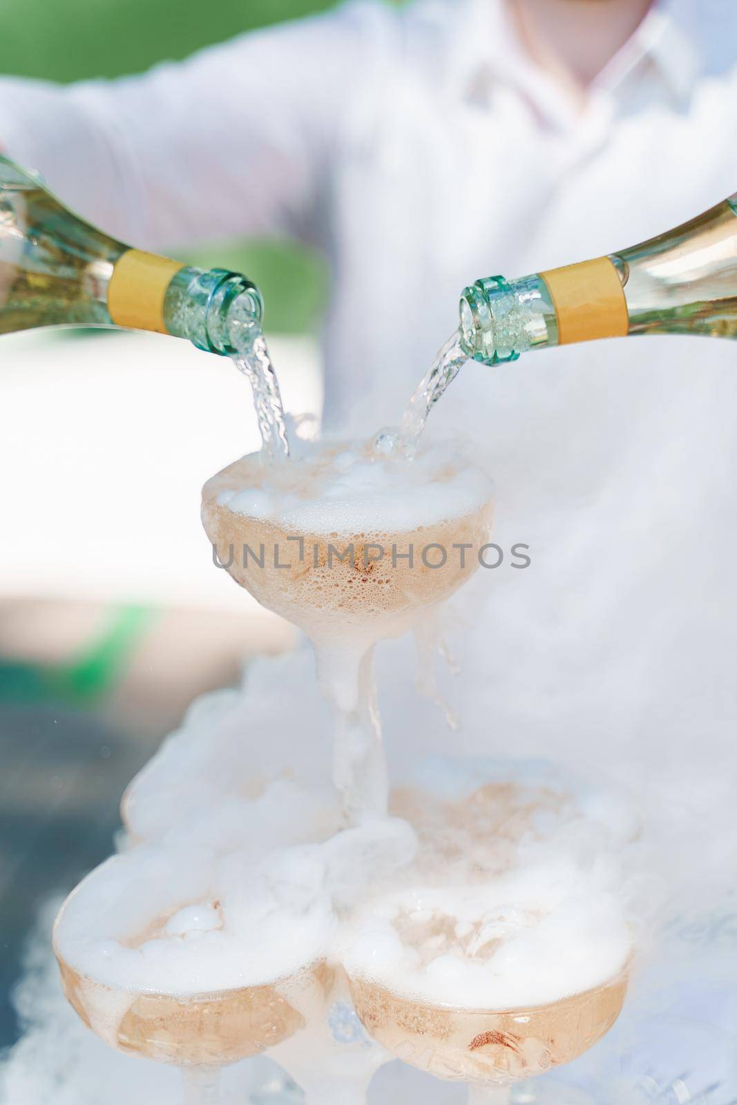Waiter pours champagne from 2 bottles into crystal glasses with dry ice and white smoke close up. Catering for business people. Vertical photo.