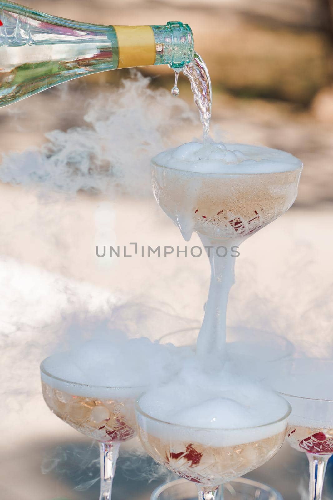 The waiter pours champagne into crystal glasses with dry ice and white smoke close up. Sparkling wine pouring in wine glass at summer day.