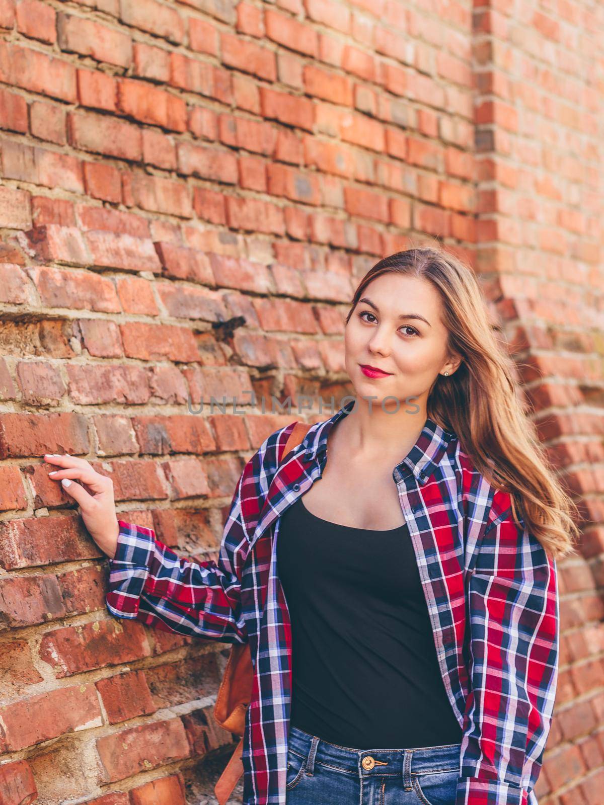 Portrait of young woman in checkered shirt and blue jeans standing against brick wall