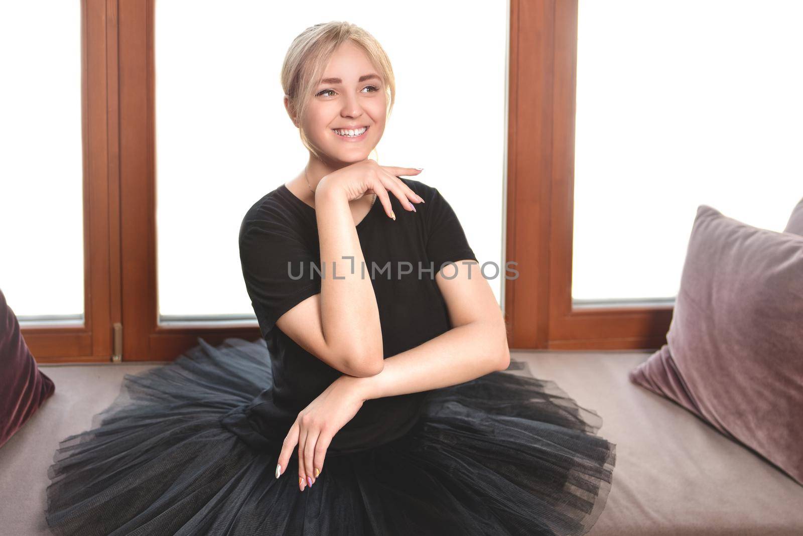 smiling ballerina in a black tutu sits in front of the window with her chin tucked in by Nickstock