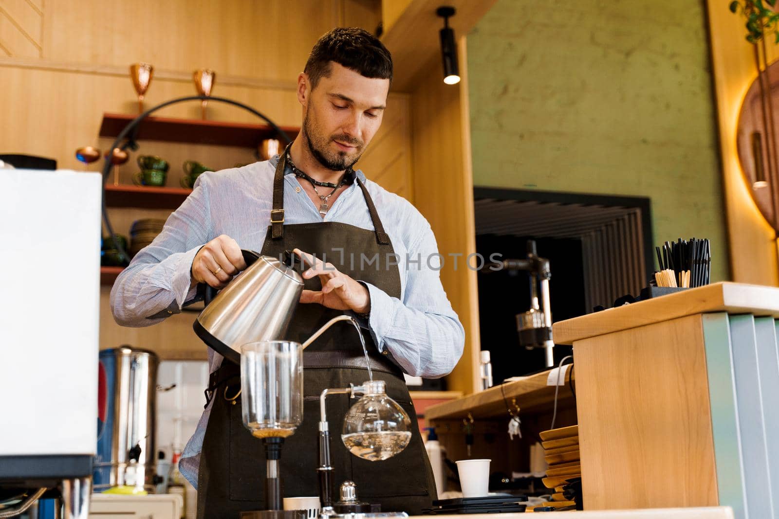 Handsome bearded barista pours hot boiling water from kattle to syphon device for coffee brewing in cafe. Syphon alternative method of making coffee.