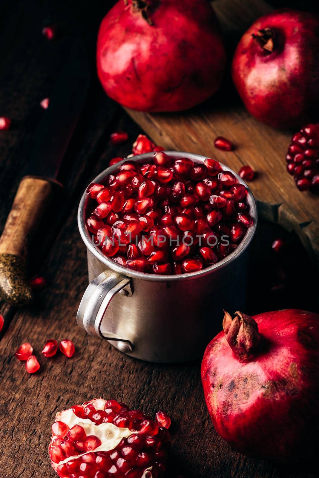 Metal rustic mug full of pomegranate seeds. Whole fruits and pomegranate pieces on dark wooden surface.