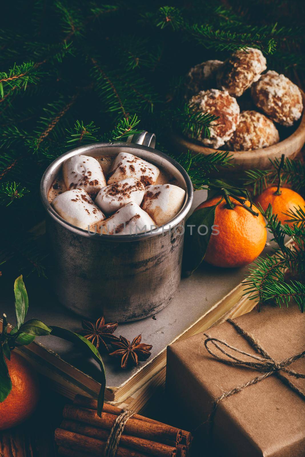 Metal mug of hot chocolate with marshmallows on stack of old books with spices and fruits