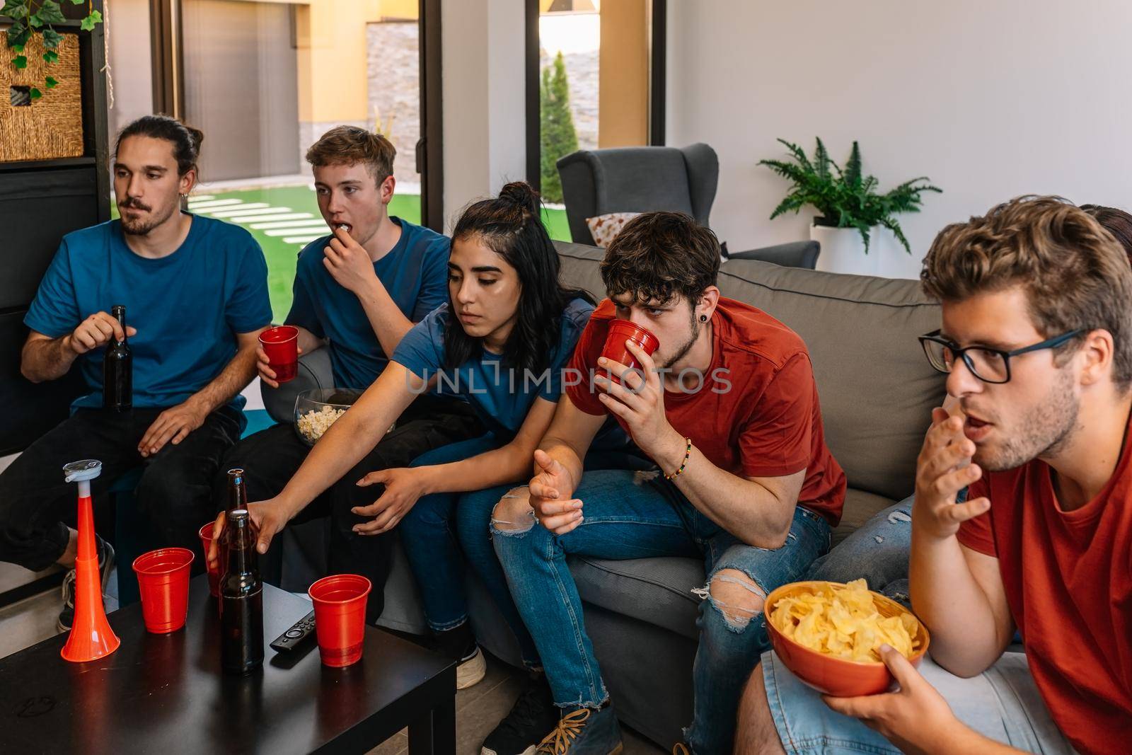 friends party at home watching television. group watching a movie or sporting event. young people eating and drinking. sitting on the sofa in the living room, natural light. table with drink and food.