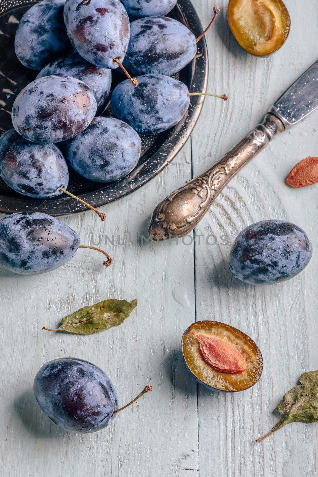 Ripe plums with sliced fruits, leaves and vintage knife over light wooden surface. View from above