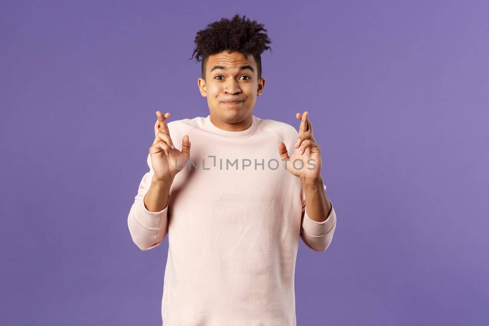 Portrait of young hopeful handsome man with dreads ancitipating something good happen, cross fingers good luck, smiling with joy and optimism, having faith, praying over purple background.