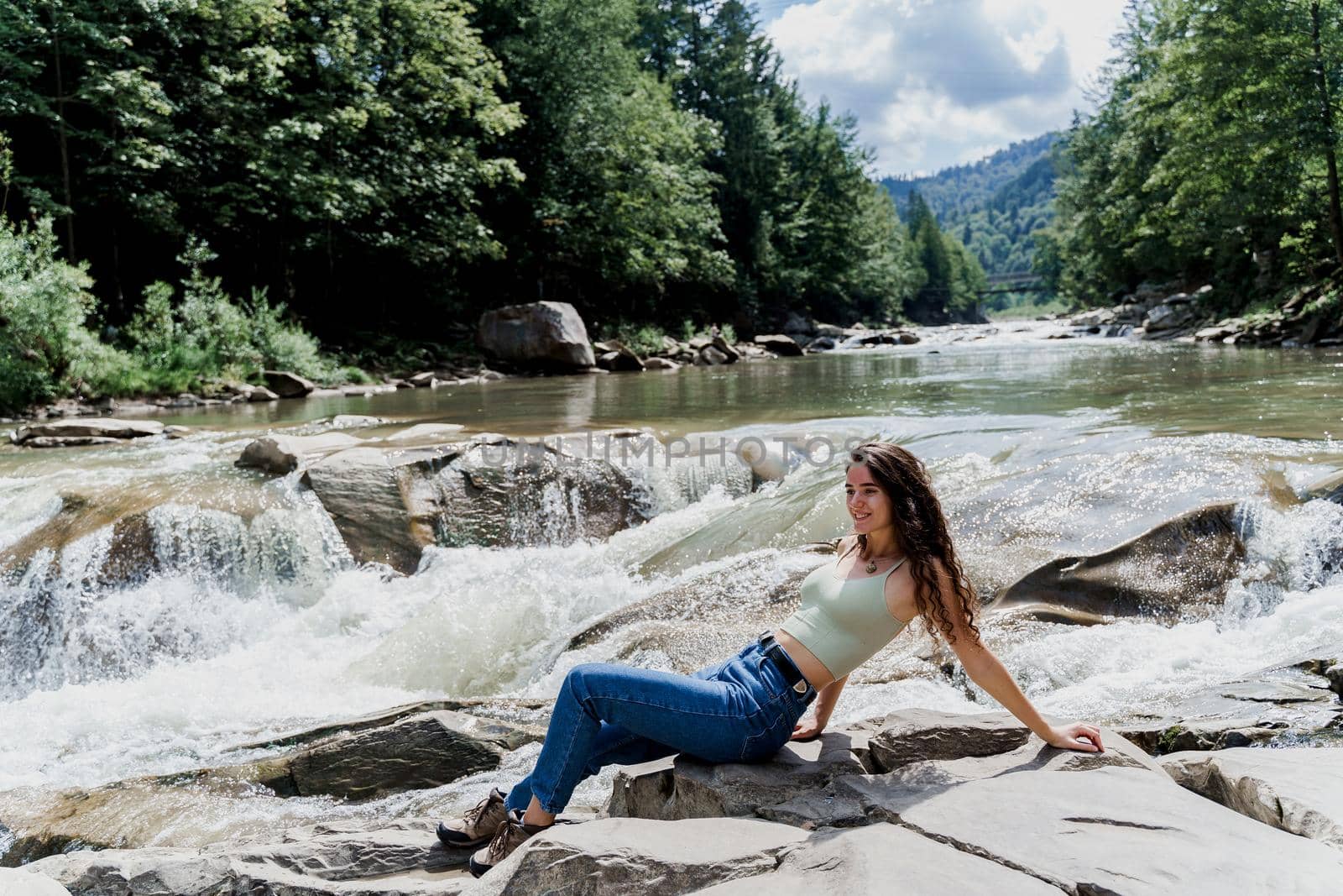Traveler girl is sitting on the rock near waterfall and looking toward. Travelling in Karpathian mountains. Cascade waterfall. Beautiful landscape