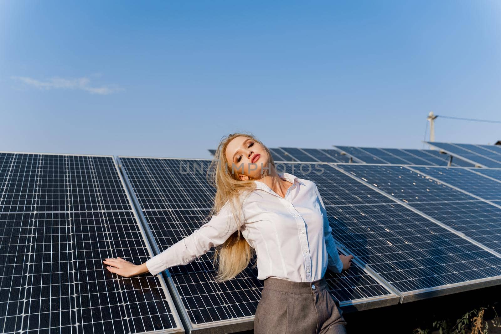 Woman leans on solar panels. Blonde dressed white formal shirt on the power plant. Free electricity for home. Green energy.