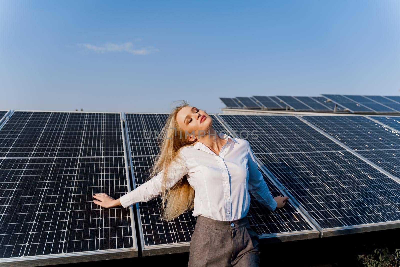 Woman leans on solar panels. Blonde dressed white formal shirt on the power plant. Free electricity for home. Green energy.