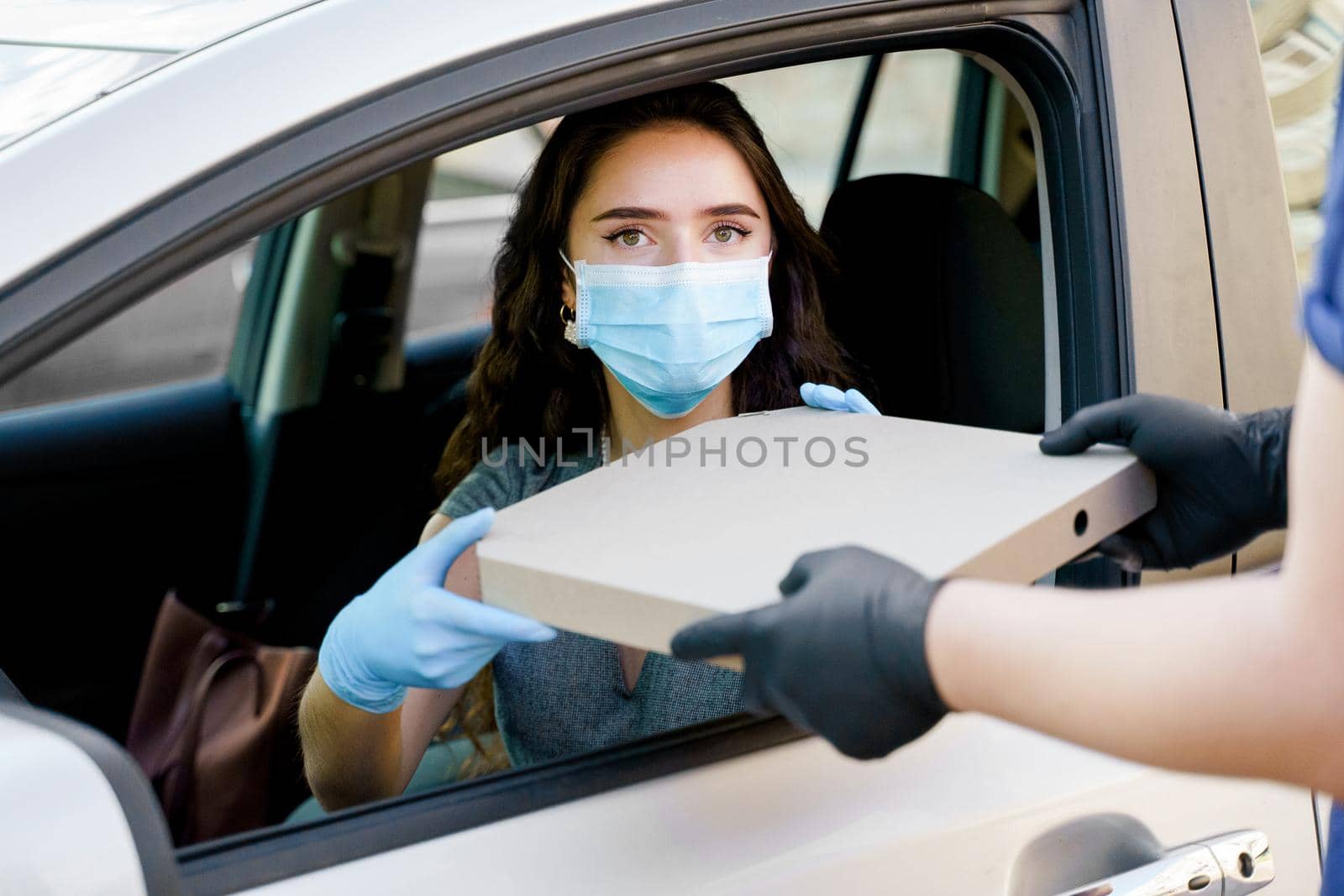 Safe food delivery from pizzeria to car during quarantine covid-19. Attractive young girl in blue medical mask and gloves gets pizza in cardboard box. by Rabizo