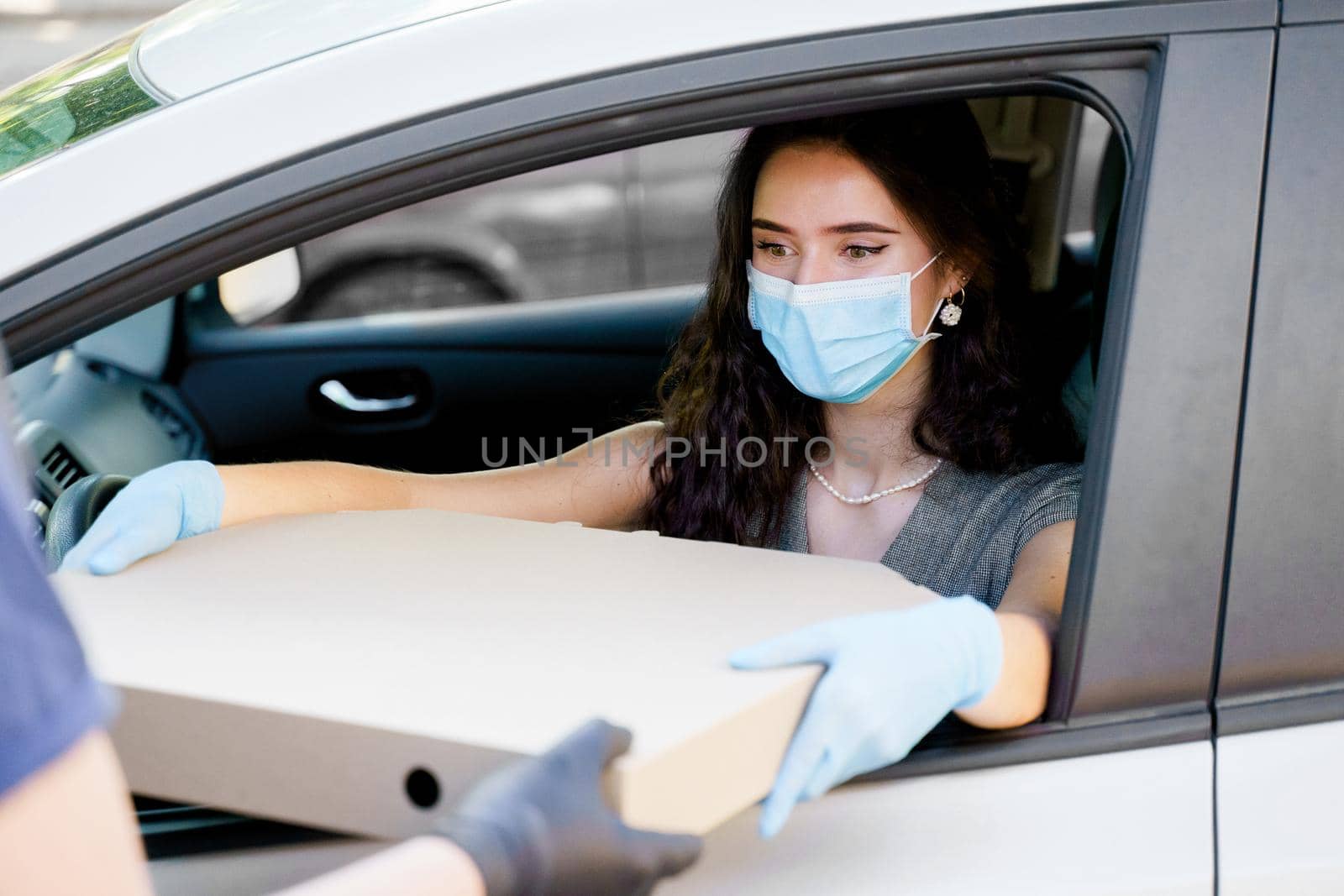 Girl in medical mask and gloves gets huge pizza in car. Big size pizza in carton eco box. Courier gives large pizza to young business woman. Delivery by car. by Rabizo