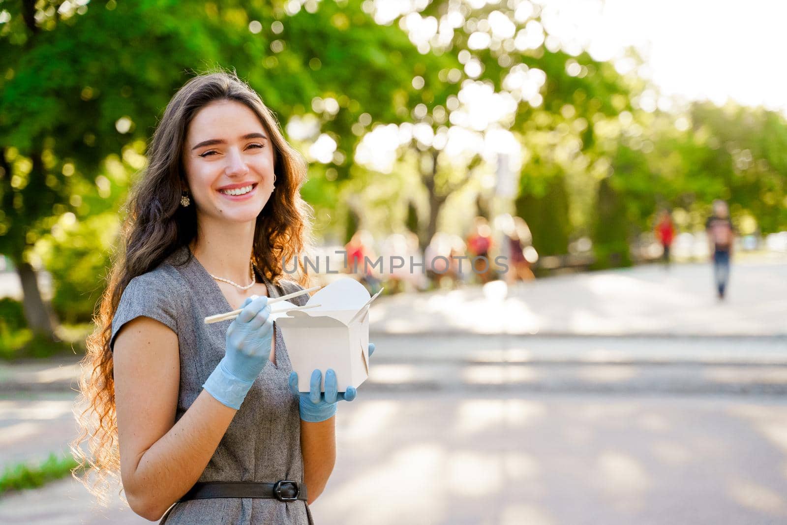 Young chinese girl with curly hair in medical gloves and mask holds wok in box in hands and smiles. Udon noodles in white box delivery